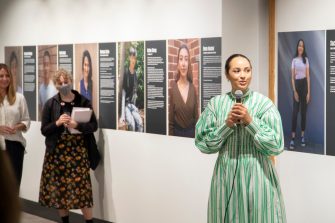 A woman wearing a striped green dress holds a microphone while giving a speech at an exhibition opening