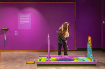 A room with purple walls and a woman with long hair in black clothing playing miniature golf.