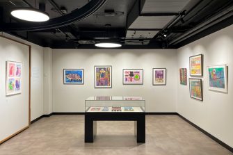 A room with white walls filled with colourful artworks in frames and a glass display case in the centre of the floor.