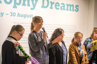 Speaker and artists standing in front of exhibition information wall, speaking to unseen crowd. Artist Emily Crockford is holding her artwork. 