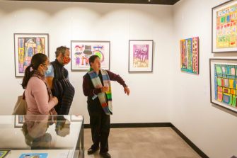 Artist in colourful striped scarf showing two guests their artwork on the wall among other colourful works.