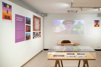 Exhibition space with main wall, display cabinets and projected video of an Indigenous person speaking.
