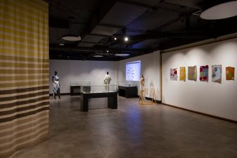 Installation view of a gallery space showing textile works, display cabinets, mannequins, and a video projection. 