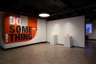 Title wall of exhibition space with a bright orange, black and white text design. Two artworks sit on white plinths next to the title wall – a large, white 3D printed coral-like object on the left, and a small, translucent orange handbag to the right.