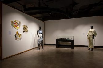 Gallery space with several artworks. Artworks include three wall-mounted handmade textile works, a mannequin with a recycled denim outfit, a display case with a black and white textile work, and a mannequin with a cream-coloured, fabric-woven outfit.