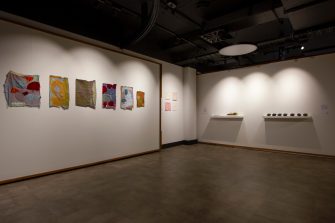 A gallery space with four separate artworks - six colourful wall-mounted textile works, four smaller red and white wall-mounted textile works, a shelf with seedling cards and sprouting greens, and a shelf with seven small, dark heart-shaped sculptures.