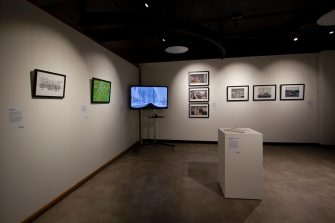Gallery space with several artworks. Artworks include a framed and wall-mounted illustration, collage, and several photographic works, a video work on a flat panel screen, and a ceramic work on a plinth. 