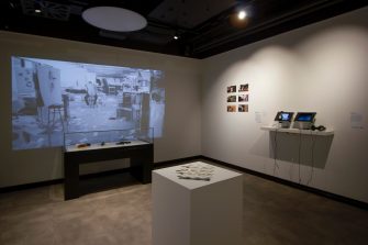 A gallery space with four separate artworks. Artworks include a video projection with a display cabinet, ceramics on a plinth, printed photographs mounted on the wall, and two iPads with headphones.