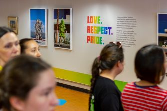 Photo of the opening event. The photo focuses on a distant wall with the exhibition title text written in colourful letters, with several framed artworks on either side. In the foreground five people view the works.