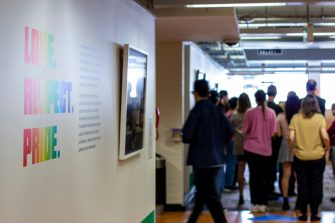 Photo of the opening event. The photo focuses on the near wall with the exhibition title text in colourful letters, with a crowd of people gathered in the background.