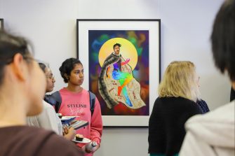 Photo of a crowd of people in front of a large, framed artwork hanging on the wall. The artwork depicts a young man with multicoloured body, wearing a fur cape, set against a bright yellow full moon.