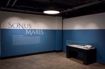 Photo of two gallery walls painted a sea-blue colour. The left wall displays the text “Sonus Maris” in large letters, with smaller, indiscernible text below. Against the right wall is a display cabinet, containing a large book and a map of Australia.