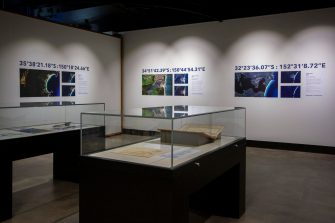 Photo of an exhibition space with two display cases in the foreground containing indiscernible items. The focus of the photo is on the walls in the background that display a series of satellite images of coastlines.