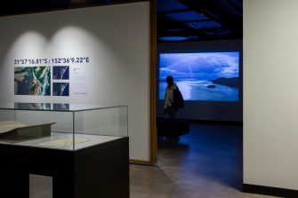 A wide-angle photo of an exhibition space. The focus is on a darkened room where a video of a coastline is being projected onto the far wall. A young woman wearing a backpack is silhouetted against the projection, watching the video.