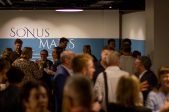 Photo of a crowd of people socialising at the exhibition opening event. In the background is a blue-and-white wall with large text reading “Sonus Maris”.