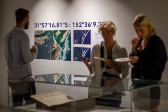 Photo of two woman holding plates, looking into a glass display cabinet. In the background, a man is looking at the gallery wall displaying satellite images of a coastline.