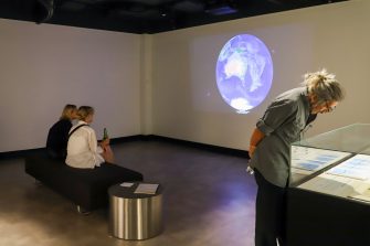 A man leaning over a display case containing documents. In the background a video is being projected onto the wall, showing an animation of the Earth as a blue globe in space. In the far left, two ladies sit on a bench, watching the video