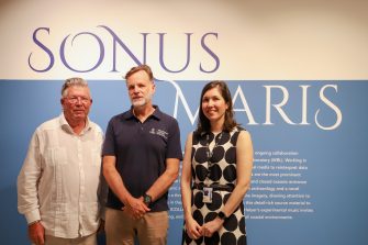 Two men and one woman standing in front of a blue and white wall with the text “Sonus Maris”, smiling at the camera.