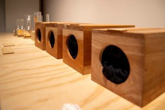 Four wooden boxes sit on a plywood table with round holes cut in the front for visitors to insert their hands and feel the contents
