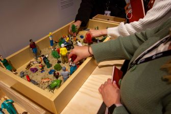Hands reach into a wooden sandbox to position figurines, plastic plants, and animals in various arrangements