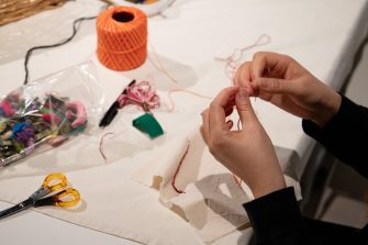 Hands carefully thread a needle with red string. On the table sits colourful thread, scissors, and calico fabric.