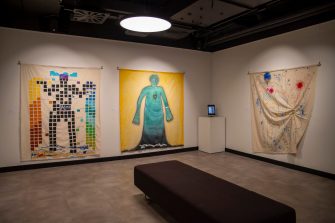 Three suspended fabric panels line two walls. On left, a figure is composed of small blocks. In the middle, a green figure stands silhouetted against a yellow background. On the right is a scrunched up fabric panel with a figurative outline. 