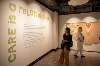 A woman stands reading wall text with the words ‘Care is a relationship’ in green block letters. A woman with her back to the camera looks at a textile artwork suspended on the wall.