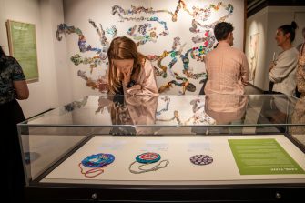 A woman bends down to look at a display case containing colourful fabric rosettes and a green paper bearing text in white letters.