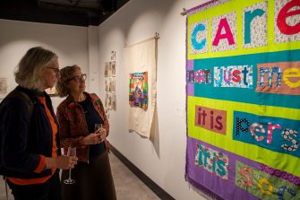 Two people stand looking at a colourful suspended artwork that features the words ' Care is not just medical it is personal it is social'