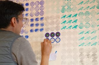 A man paints a wall with a fine paint brush. On the wall is a repeating pattern of concentric circles in blue, black, and green ink. Patterns of birds and flowers are also visible. 