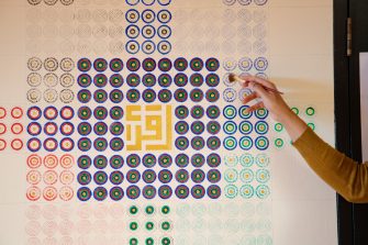 Photo of an elaborate painting on a wall featuring a square pattern made up of individual circles in a grid formation. The circles are painted an alternating pattern of red, green, blue and black. In the centre of the square pattern is a gilded gold kufic calligraphy symbol. 