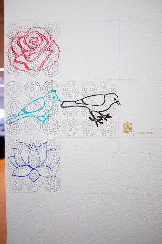 A white wall with four striking symbols created with a woodblock stamp: a rose, a lotus flower, a nightingale, and a myna bird. To the right of the myna bird print is a small gold signature.
