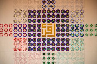 Photo of an elaborate painting on a wall featuring a square pattern made up of individual circles in a grid formation. The circles are painted an alternating pattern of red, green, blue and black. In the centre of the square pattern is a gilded gold Kufic calligraphy emblem. 