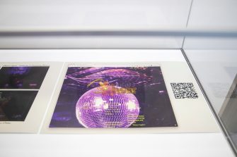 A close-up of a grey poster lying in a glass cabinet. The poster displays an image of a purple disco-ball.