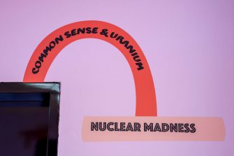 A purple wall with an illustration of two book spines. The titles of the books read "Common Sense & Uranium" and "Nuclear Madness". 