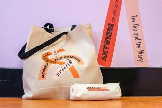 A pencil case and a tote bag on the ground next to a corner of the mural wall. The merchandises and mural have the same designs: curvy orange lines with text in different fonts on top of each line.