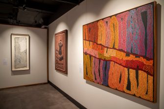 A gallery with three beautiful paintings handing on the wall. The closest painting to the camera depicts abstracted patterns in bright orange, purple, and brown colours. 