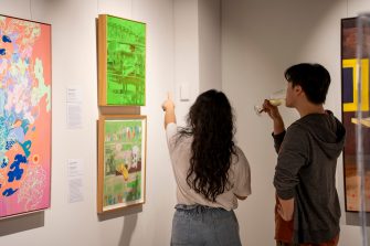 Two people stand in front of two bright green artworks hanging on the wall. One viewer points to the artwork, the other sips a glass of wine.