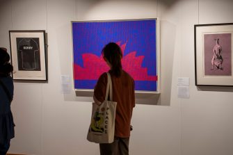 A woman stands in front of a bright blue and pink painting depicting the Sydney Opera House.