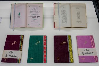 A view inside the glass display cabinet, containing six books. In the upper half of the image shows two books opened to reveal the beginning pages of each volume. The first book reads "The Mahabharata" in large ornate lettering. The lower half of the image displays four closed books sitting side-by-side. The covers are handbound in colourful fabric with decorative embroidery.