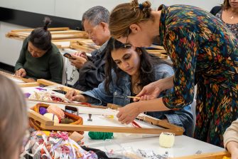 Several people sitting at a table during a weaving workshop. A woman assists another with a loom, surrounded by colourful yarns.