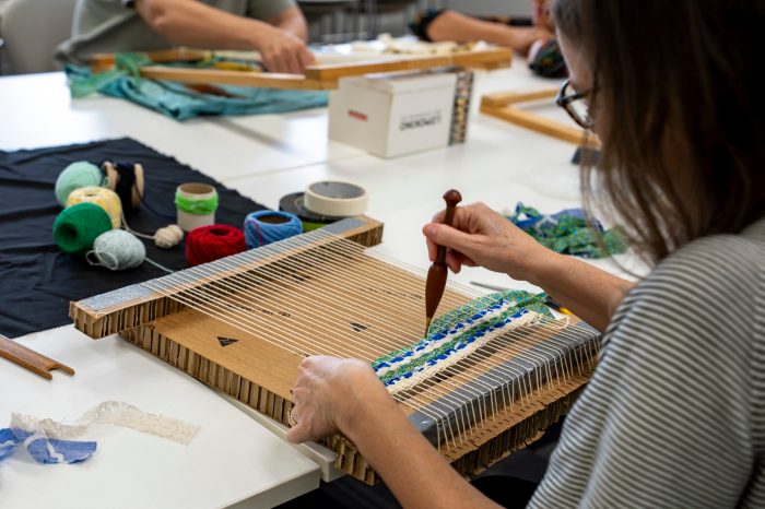 A person weaves blue and green yarn on a handmade cardboard loom. Spools of colourful yarn and tools are scattered on a white table.
