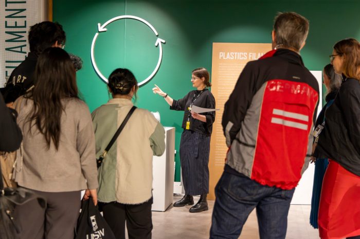 A group of people standing and listening to a woman explaining a display on recycling. Behind the woman is a circular arrow symbol is on a green wall, symbolising circular economy themes. 