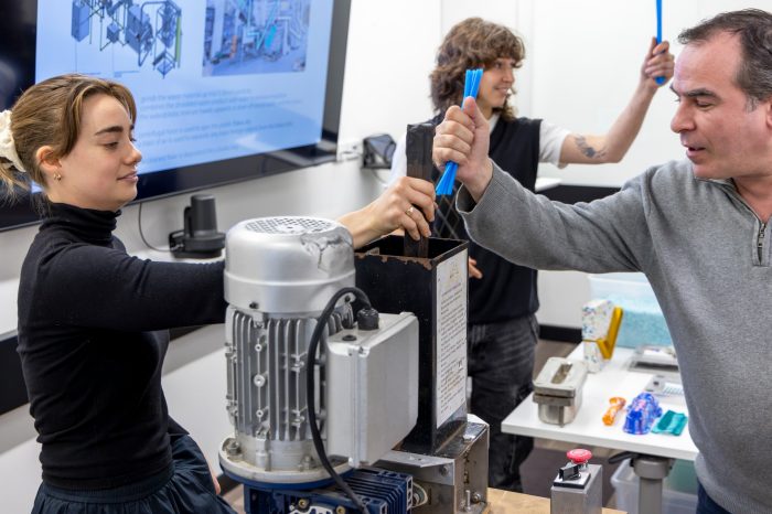 Three people stand at the front of a classroom, demonstrate inserting plastic items into recycling machinery.
