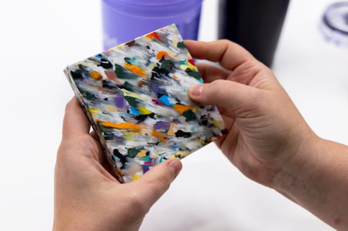 Hands holding a colourful, abstract-patterned, recycled plastic tile. 