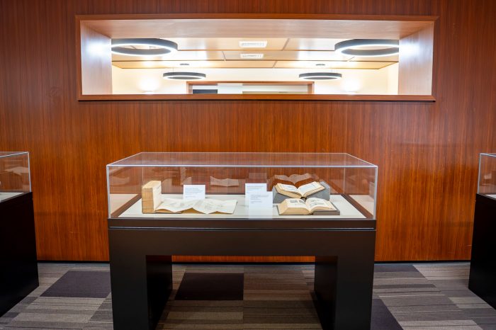 A glass display case containing several books with fold out pages. In the background is a wood veneer wall with circular fluorescent lights. 