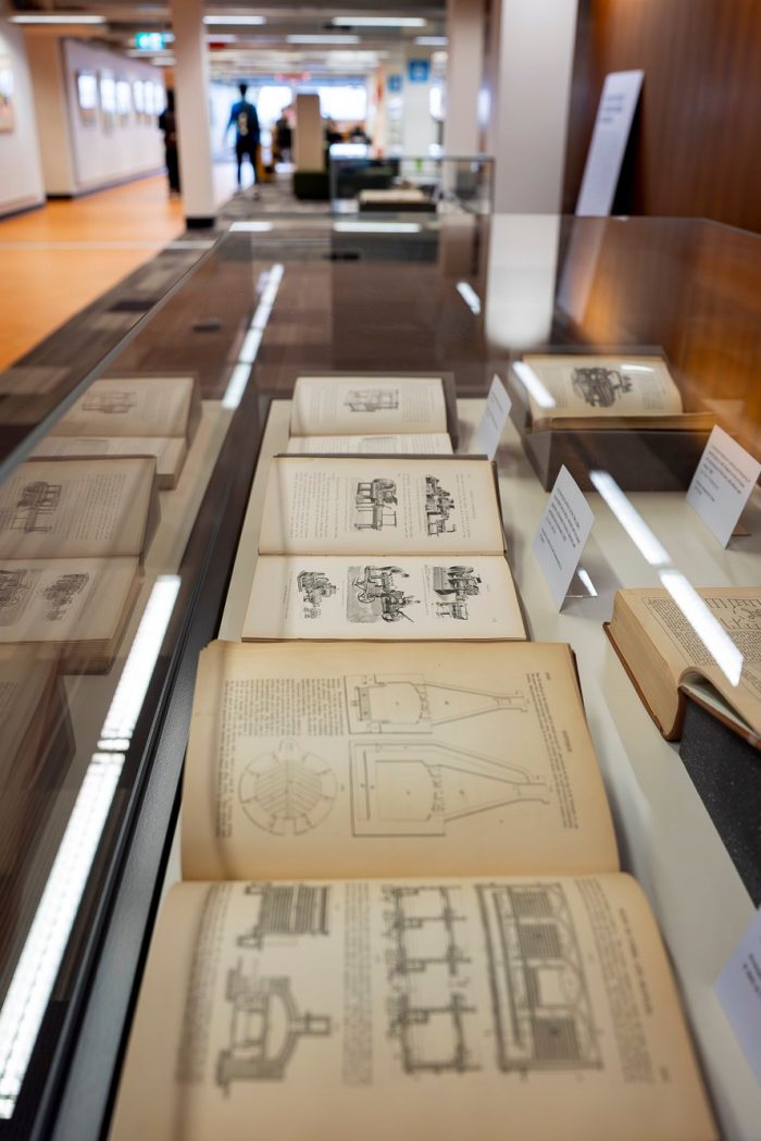 A side-on view of a glass display case containing several old books opened to pages showing descriptive illustrations of 19th Century machinery