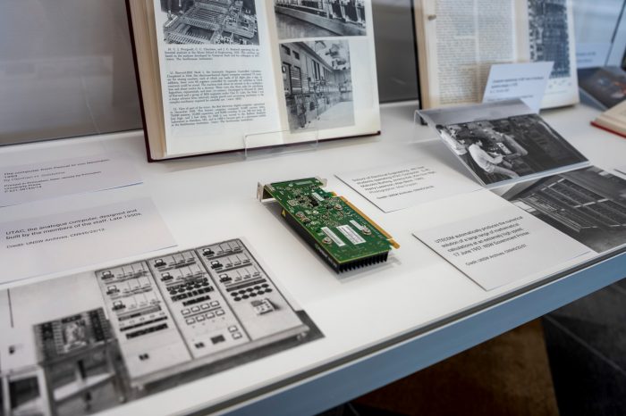 Close-up of the contents inside a glass display case. There are three black and white photos of vintage computers, and a modern computer CPU board.