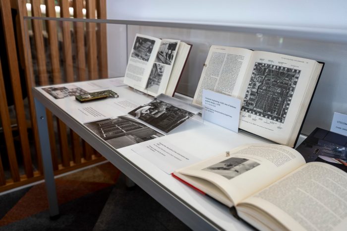 Side view of a glass display case containing several open books. The image focuses on a book showing a microscopic image of an integrated circuit inside a computer.