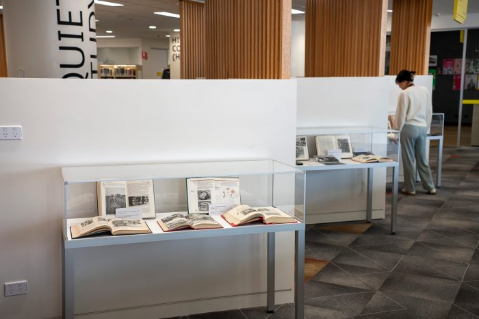 A photo of a library with three display cases in a row. Inside the display cases are several opened books. A lady wearing blue pants and a white knitted top looks into one of the display cases. 